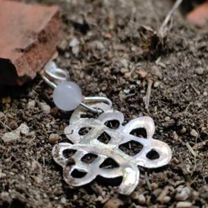 Pendentif "trèfle" celtique argent et quartz rose, posé sur de la terre de mon jardin comme un trésor qu'un archéologue ou historien aurait déterré après avoir passé des siècles sous terre. Collection Artéfacts chez Le Chêne Enchanté. Vu entière du pendentif.