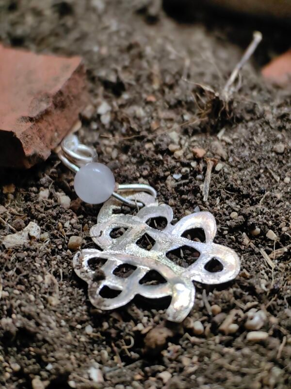 Pendentif "trèfle" celtique argent et quartz rose, posé sur de la terre de mon jardin comme un trésor qu'un archéologue ou historien aurait déterré après avoir passé des siècles sous terre. Collection Artéfacts chez Le Chêne Enchanté. Vu entière du pendentif.