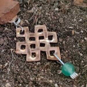 Pendentif "noeud de la longévité" en argent-cuivre éthiques et aventurine verte, posé sur de la terre de mon jardin comme un trésor qu'un historien ou archéologue aurait déterré après des siècles passés sous terre. Collection Artéfacts chez Le Chêne Enchanté. Vu entière du pendentif.