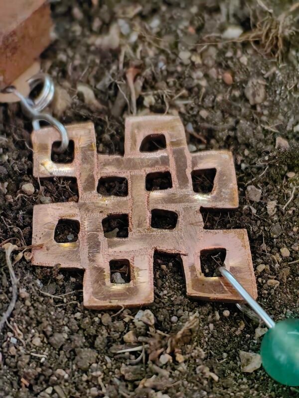 Pendentif "noeud de la longévité" en argent-cuivre éthiques et aventurine verte, posé sur de la terre de mon jardin comme un trésor qu'un historien ou archéologue aurait déterré après des siècles passés sous terre. Collection Artéfacts chez Le Chêne Enchanté. Vu sur le noeud de la longévité.