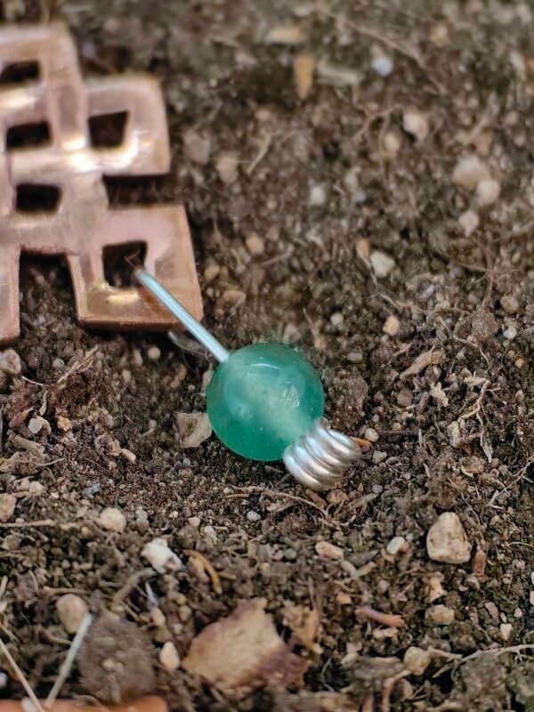 Pendentif "noeud de la longévité" en argent-cuivre éthiques et aventurine verte, posé sur de la terre de mon jardin comme un trésor qu'un historien ou archéologue aurait déterré après des siècles passés sous terre. Collection Artéfacts chez Le Chêne Enchanté. Vu sur l'aventurine verte naturelle.