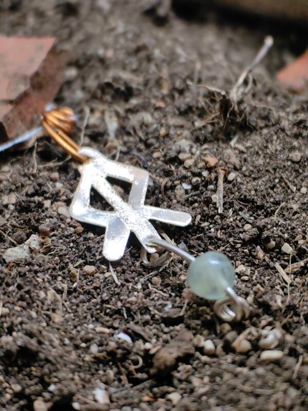 Pendentif rune courage, argent-cuivre et cristal de roche, collection artéfacts chez Le Chêne Enchanté. Le pendentif runique est délicatement déposé sur la terre de mon jardin, comme aurait pu le découvrir un archéologue ou un historien après qu'il ait été déterré après avoir passé des siècles sous terre. Vu entière du pendentif.