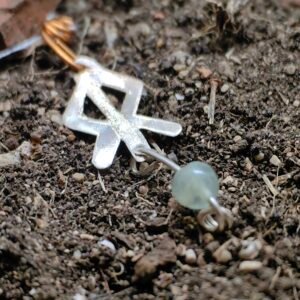 Pendentif rune courage, argent-cuivre et cristal de roche, collection artéfacts chez Le Chêne Enchanté. Le pendentif runique est délicatement déposé sur la terre de mon jardin, comme aurait pu le découvrir un archéologue ou un historien après qu'il ait été déterré après avoir passé des siècles sous terre. Vu entière du pendentif.