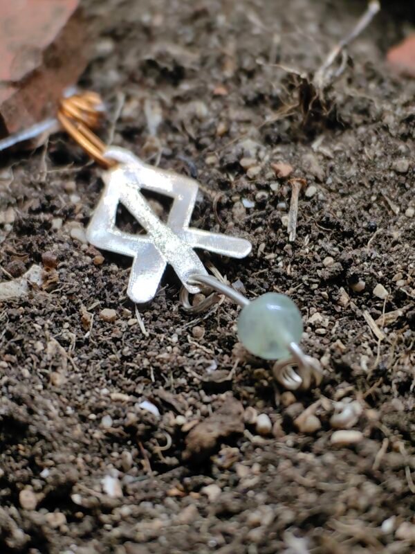 Pendentif rune courage, argent-cuivre et cristal de roche, collection artéfacts chez Le Chêne Enchanté. Le pendentif runique est délicatement déposé sur la terre de mon jardin, comme aurait pu le découvrir un archéologue ou un historien après qu'il ait été déterré après avoir passé des siècles sous terre. Vu entière du pendentif.