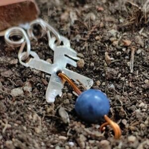 Pendentif rune chance en argent-cuivre étiques et merlinite, collection artéfact chez Le Chêne Enchanté. Le pendentif runique est délicatement posé sur la terre de mon jardin comme aurait pu le découvrir un archéologue ou un historien des siècles après qu'il ait été enterré. Vu entière du pendentif.