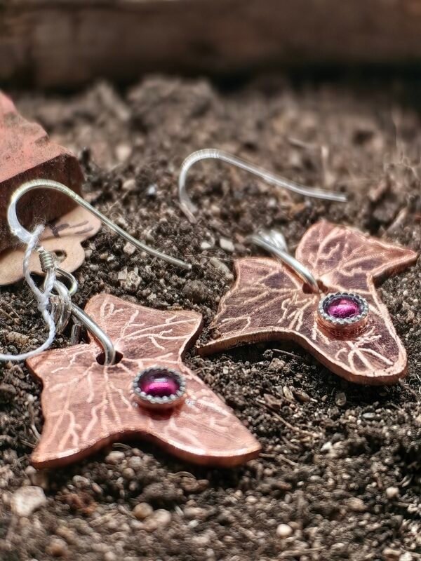 Boucles d'oreilles lierre mystique, en argent-cuivre éthiques et grenats éthiques, collection artéfacts chez Le Chêne Enchanté. Les boucles d'oreilles sont posées sur la terre de mon jardin comme l'aurait découvert un archéologue ou un historien après avoir passé des siècles enterré. Vu entière des boucles d'oreilles.