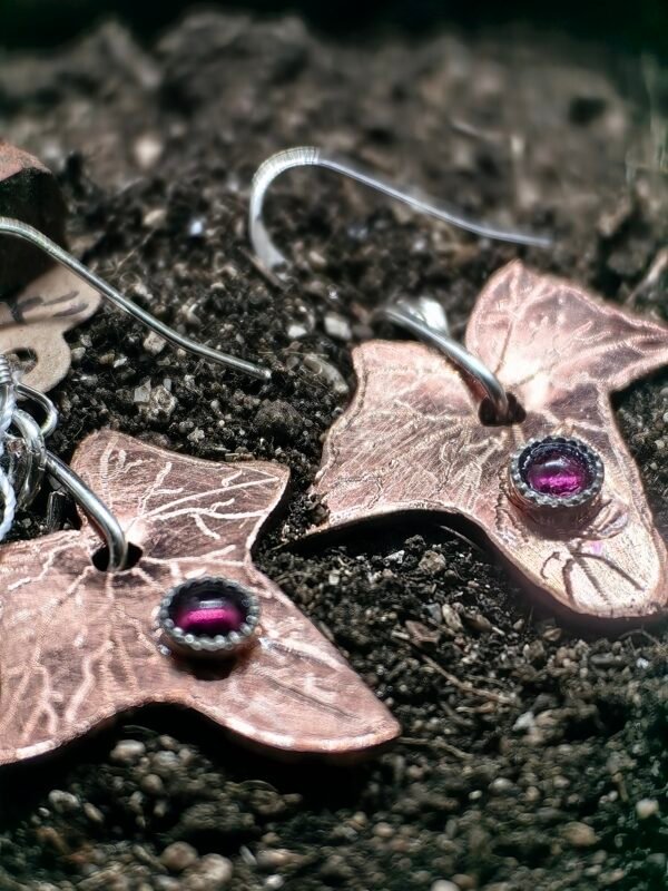 Boucles d'oreilles lierre mystique, en argent-cuivre éthiques et grenats éthiques, collection artéfacts chez Le Chêne Enchanté. Les boucles d'oreilles sont posées sur la terre de mon jardin comme l'aurait découvert un archéologue ou un historien après avoir passé des siècles enterré. Vu de près des boucles d'oreilles avec un gros plan sur les grenats sertis d'argent.