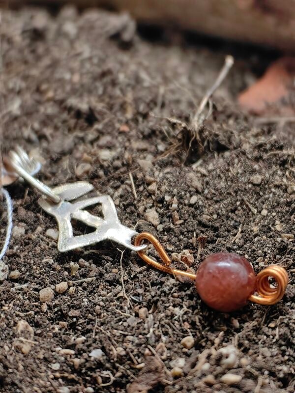Pendentif rune protection en argent-cuvire et cornaline, collection artéfacts chez Le Chêne Enchanté. Le pendentif est posé sur la terre de mon jardin comme l'aurait découvert des archéologues et historiens après avoir passé des siècles enterrés sous terre. Vu entière du pendentif.