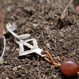 Pendentif rune protection en argent-cuvire et cornaline, collection artéfacts chez Le Chêne Enchanté. Le pendentif est posé sur la terre de mon jardin comme l'aurait découvert des archéologues et historiens après avoir passé des siècles enterrés sous terre. Vu de près du pendentif.