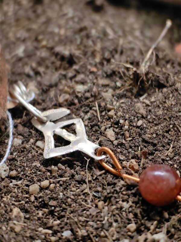 Pendentif rune protection en argent-cuvire et cornaline, collection artéfacts chez Le Chêne Enchanté. Le pendentif est posé sur la terre de mon jardin comme l'aurait découvert des archéologues et historiens après avoir passé des siècles enterrés sous terre. Vu de près du pendentif.