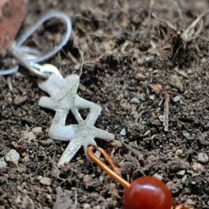 Pendentf rune protection en argent-cuivre et cornaline, collection Artéfacts chez Le Chêne Enchanté. Le pendentif est posé sur la terre de mon jardin comme un ancien trésor qu'un archéologue ou un historien aurait découvert des siècles après avoir été enterré. Vu entière du pendentif.
