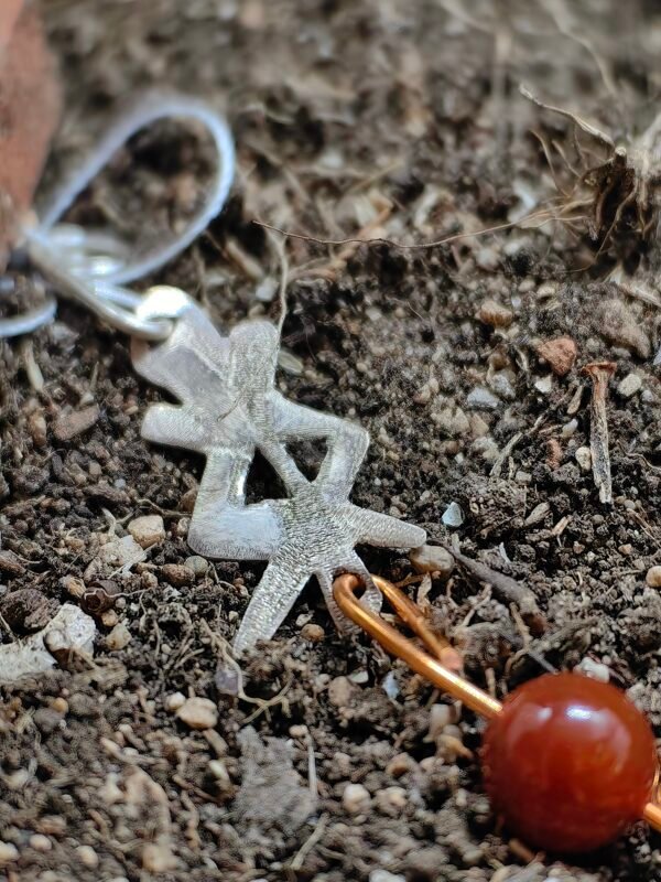 Pendentif rune protection en argent-cuivre et cornaline, collection Artéfacts chez Le Chêne Enchanté. Le pendentif est posé sur la terre de mon jardin comme un ancien trésor qu'un archéologue ou un historien aurait découvert des siècles après avoir été enterré. Vu de près du pendentif.