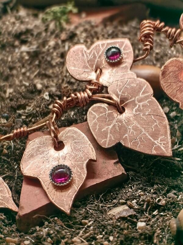 Gros plans sur les feuilles de lierres du collier "lierre mystique" argent-cuivre éthiques et grenat, collection artéfacts chez Le Chêne Enchanté. Le collier est posé sur la terre de mon jardin, certaines de ses feuilles de lierres sont partiellement enterrées comme l'aurait découvert un archéologue ou un histoirien après que le collier ait passé des siècles sous terre.