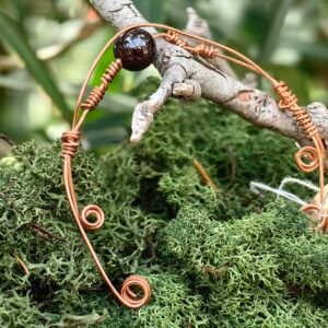 Bracelet Force intérieure en fils de cuivre tressés et grenat en perle, collection Artéfacts chez Le Chêne Enchanté. Le bracelet incarne les forces telluriques, l'amour et la protection. Ce bracelet réglable sous forme de jonc souple repose sur une branche de chêne au milieu d'une mousse forestière verte naturelle. Le bracelet se termine par deux spirales à chaque de ses extrêmités. Tous les bijoux et photographies sont l'oeuvre de : Le Chêne Enchanté et interdite de reproduction.