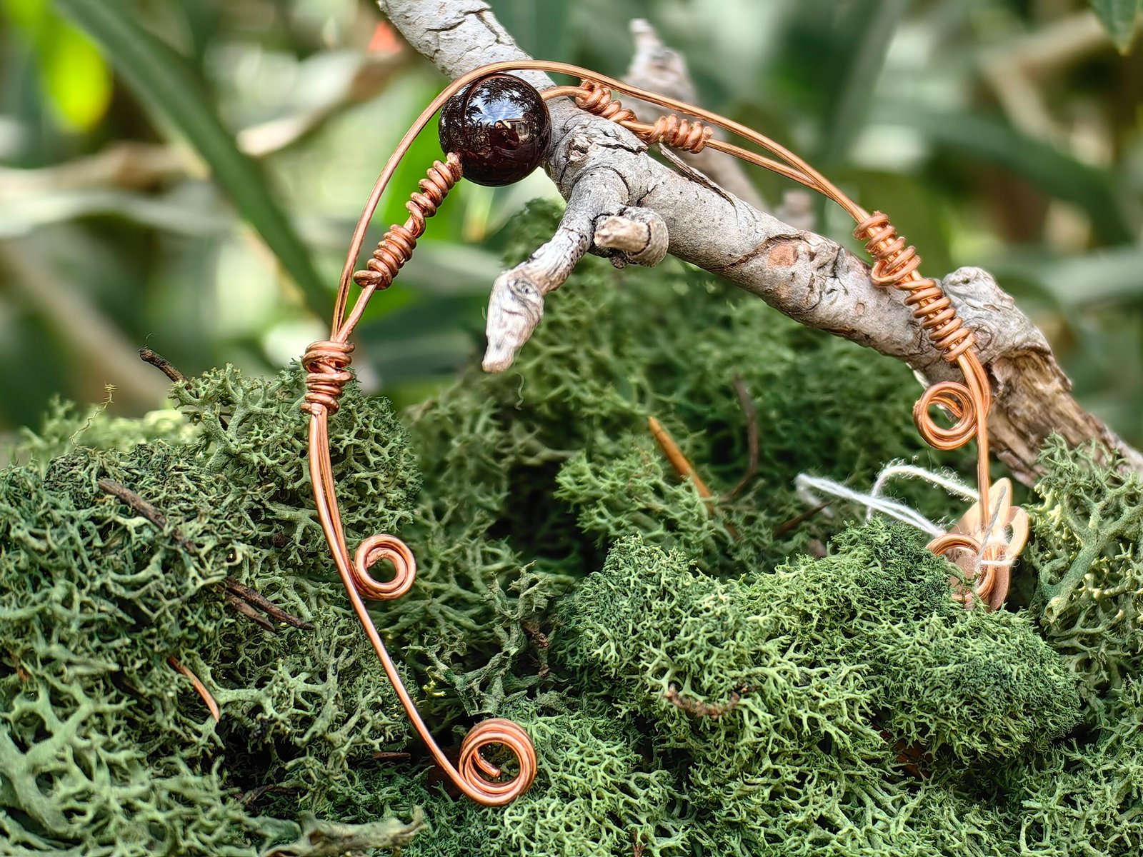 Bracelet Force intérieure en fils de cuivre tressés et grenat en perle, collection Artéfacts chez Le Chêne Enchanté. Le bracelet incarne les forces telluriques, l'amour et la protection. Ce bracelet réglable sous forme de jonc souple repose sur une branche de chêne au milieu d'une mousse forestière verte naturelle. Le bracelet se termine par deux spirales à chaque de ses extrêmités. Tous les bijoux et photographies sont l'oeuvre de : Le Chêne Enchanté et interdite de reproduction.