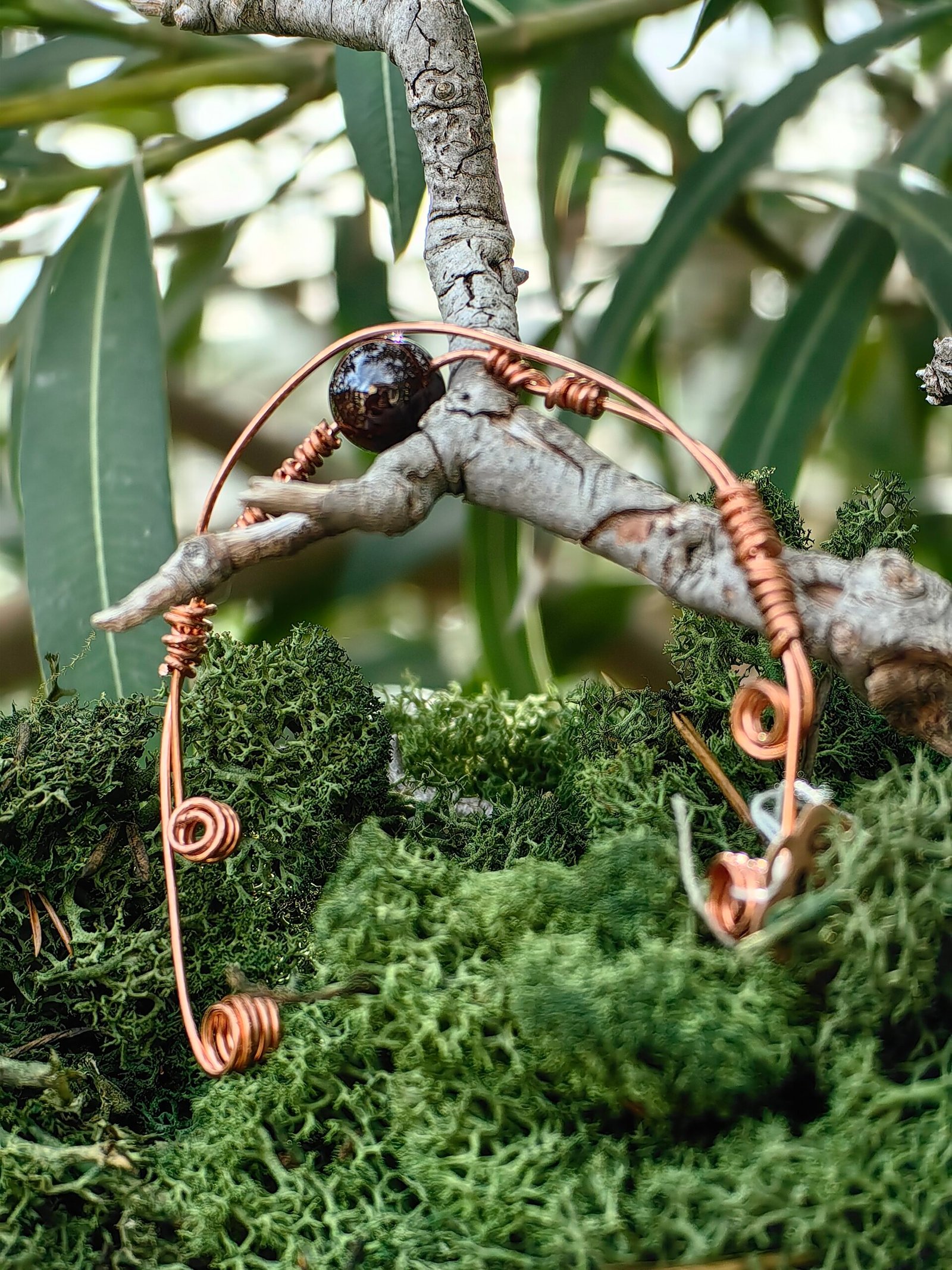 Bracelet Force intérieure en fils de cuivre tressés et grenat en perle, collection Artéfacts chez Le Chêne Enchanté. Le bracelet incarne les forces telluriques, l'amour et la protection. Ce bracelet réglable sous forme de jonc souple repose sur une branche de chêne au milieu d'une mousse forestière verte naturelle. Le bracelet se termine par deux spirales à chaque de ses extrêmités. Vu de côté du bracelet. Tous les bijoux et photographies sont l'oeuvre de : Le Chêne Enchanté et interdite de reproduction.